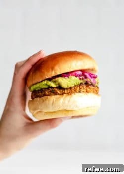 A hand holding a chickpea veggie burger filled with guacamole and pickled cabbage, presented appealingly.