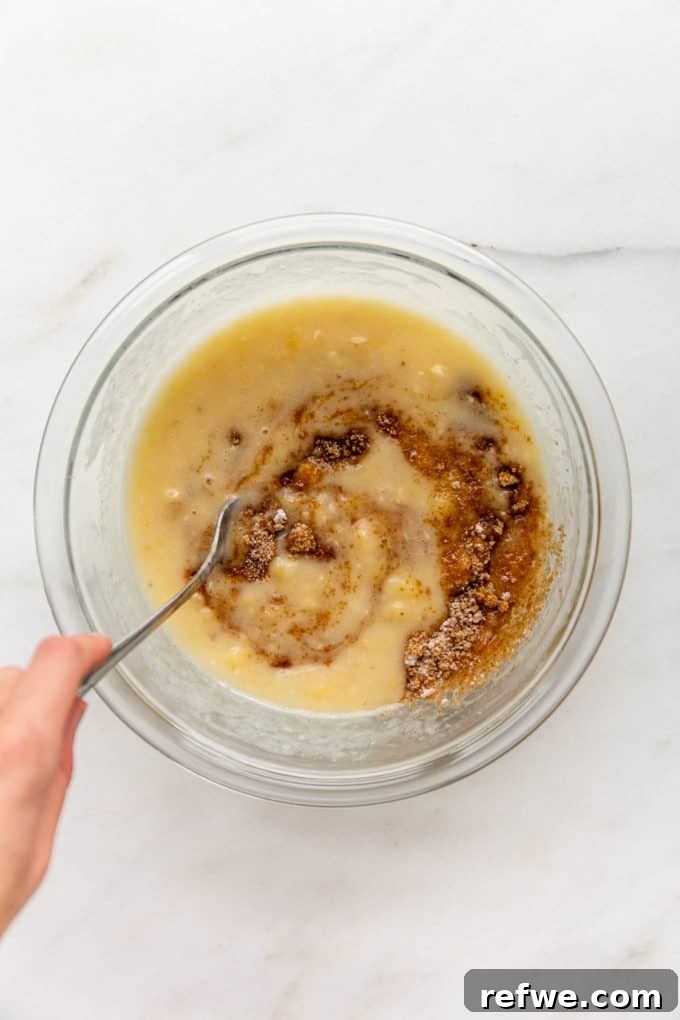 A hand mixing brown sugar into a bowl of mashed bananas and other wet ingredients.