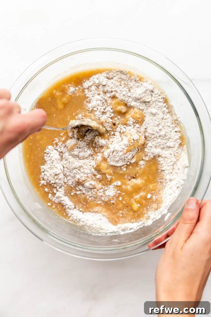 A hand gently mixing flour with bananas in a mixing bowl, illustrating the folding technique.