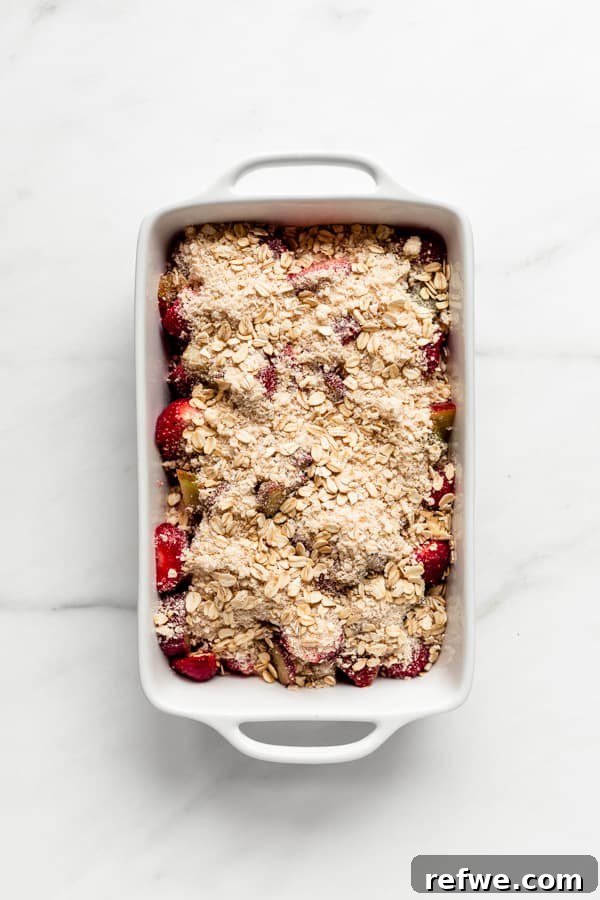 A white baking dish filled with a vibrant strawberry and rhubarb base, generously covered with a golden oat crumble topping.