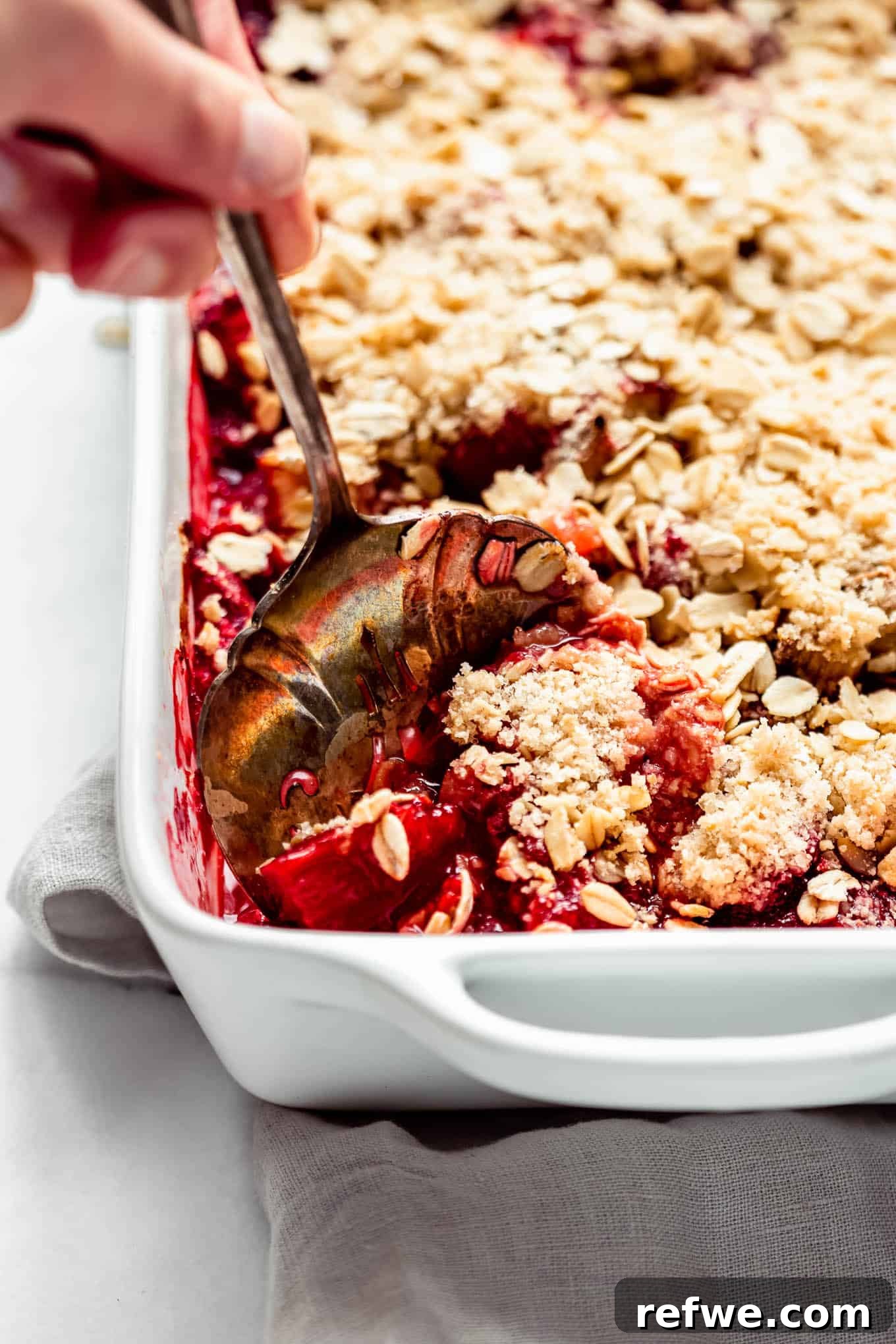 A hand scooping a generous portion of freshly baked healthy strawberry rhubarb crisp, highlighting its crispy topping and bubbly fruit filling.