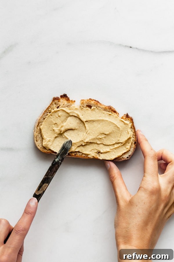 Effortless Hummus Toast Explorations 5 A close-up shot of a hand gently spreading a thick layer of smooth, light-colored hummus onto a golden-brown slice of toasted bread using a silver knife.