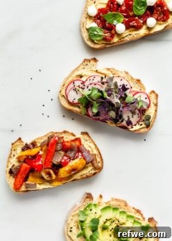 Four pieces of hummus on toast with various fresh toppings, arranged attractively on a wooden board, ready to be served.