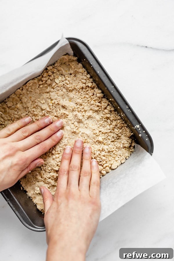 Golden Raspberry Crumble Bars 5 Two hands pressing the crumble dough mixture firmly into an 8x8 inch pan to form an even crust.