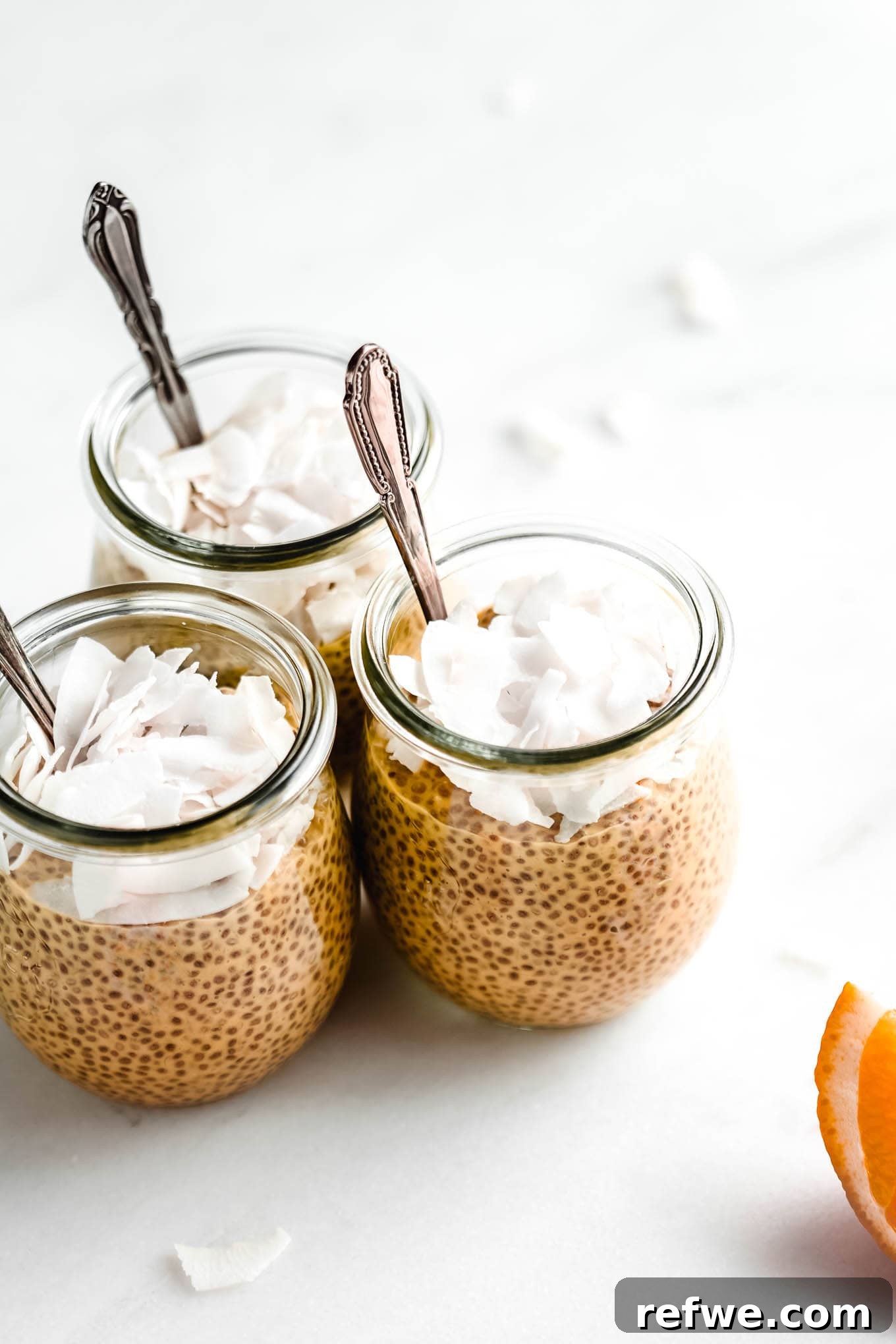 Silky Orange Dream Chia Pudding 2 Three jars of orange creamsicle chia pudding topped with shredded coconut and spoons.