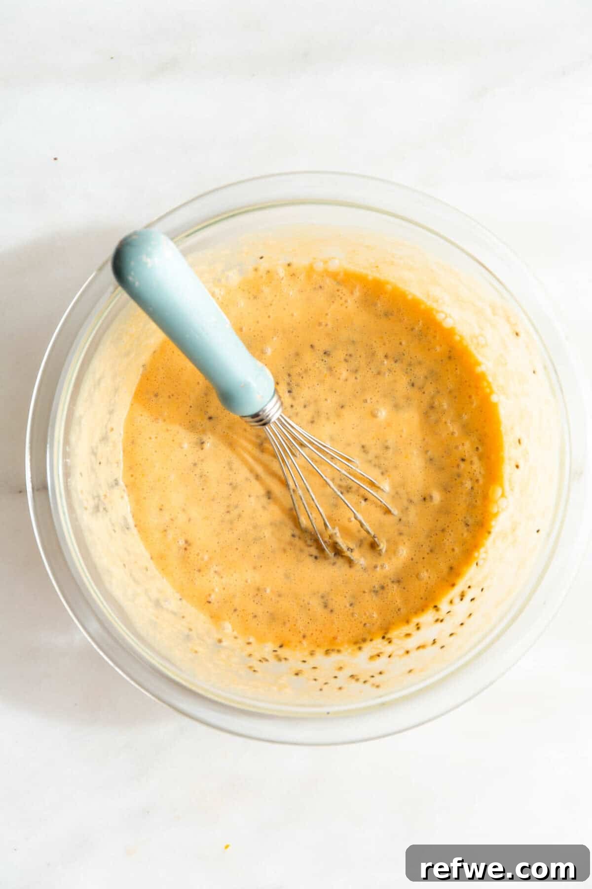 Silky Orange Dream Chia Pudding 7 The orange chia seed pudding mixture is being whisked in a clear mixing bowl with a blue whisk.