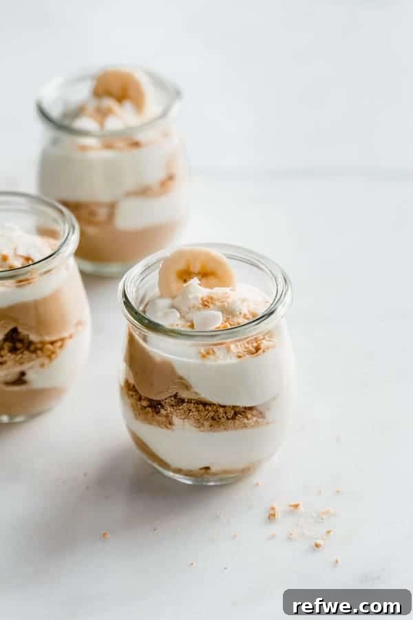 A beautifully layered cup of vegan banana cream pie parfait, showcasing rich vanilla pudding, white coconut whipped cream, golden graham cracker crumbs, and fresh banana slices