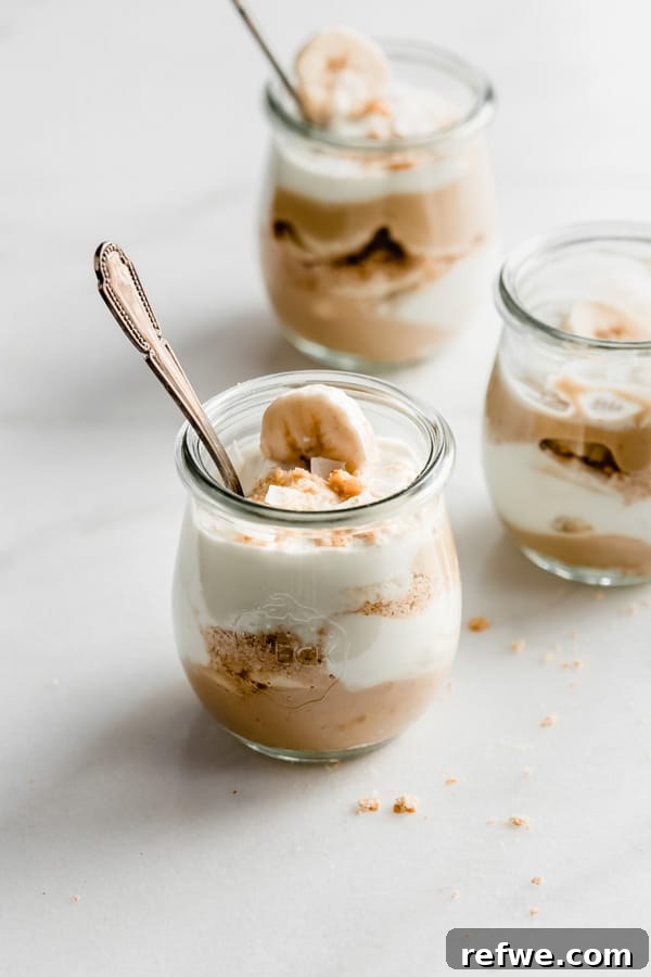Three beautifully presented cups of vegan banana cream pie parfaits, artfully garnished with additional graham cracker crumbs scattered around them for a rustic touch