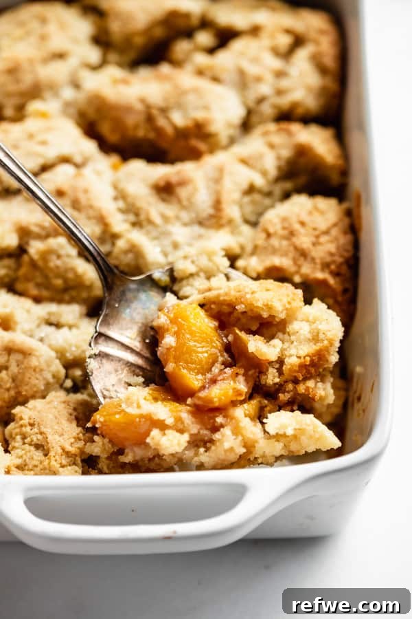Luscious Gluten-Free Peach Cobbler 6 A large serving spoon scooping a generous portion of warm, gluten-free peach cobbler from a white baking dish, ready to be enjoyed.