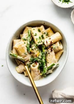 A bowl of vegan lemon asparagus rigatoni noodles garnished with fresh parsley and chili flakes, with small side bowls.