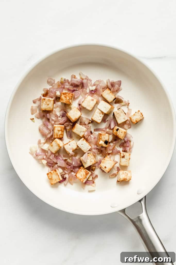 Crispy, golden-brown tofu cubes stir-frying alongside shallots in a white pan, creating a delightful texture.