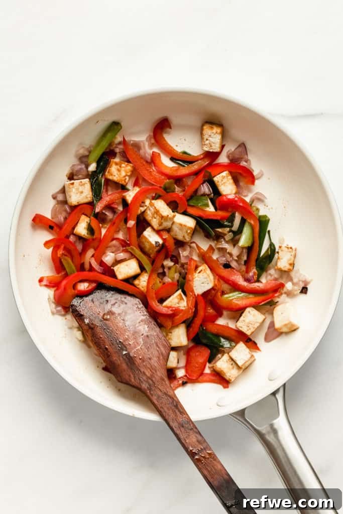 A colorful mix of crispy tofu, vibrant red bell peppers, and fresh green onions stir-frying together in a white pan.