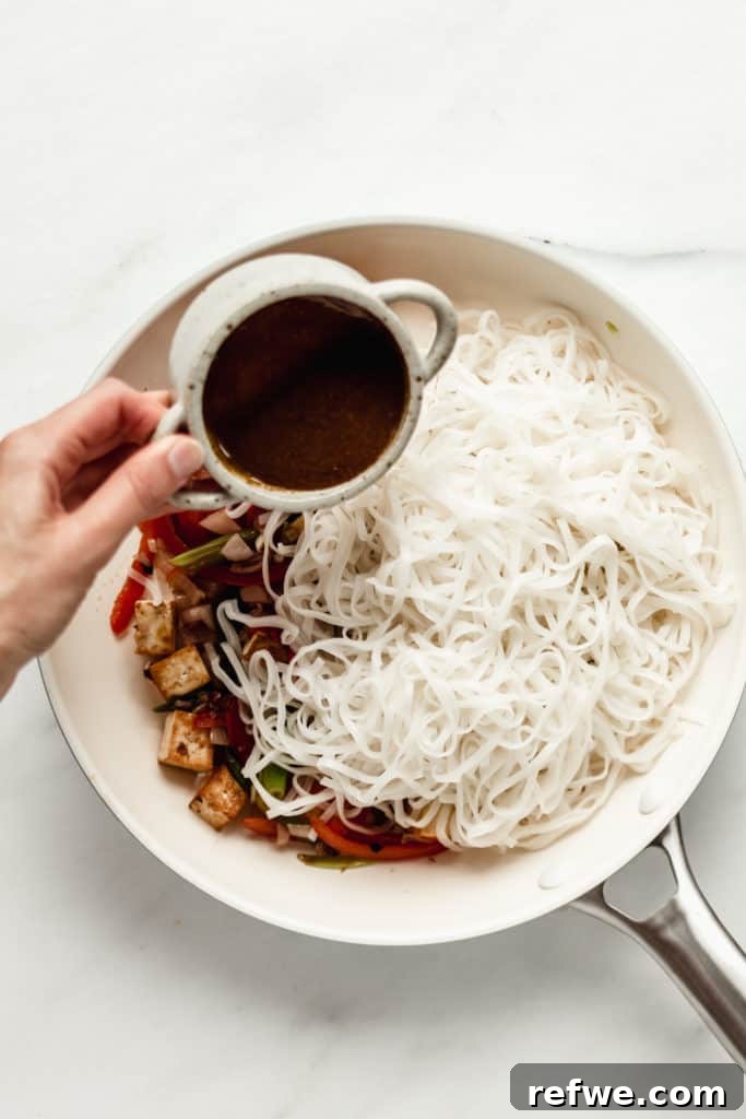 Cooked rice noodles being tossed in a pan, with a hand pouring the flavorful sauce over them to coat evenly.