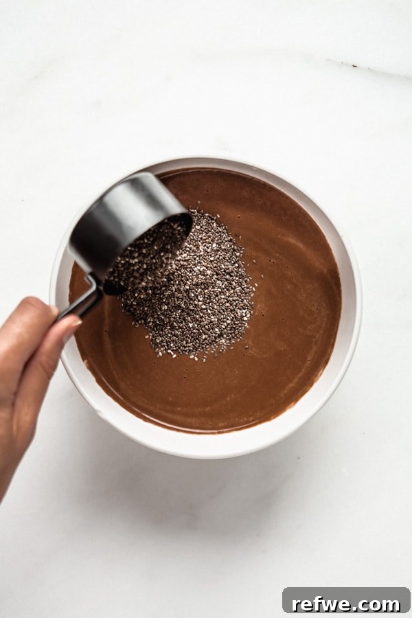 Decadent Chocolate Raspberry Chia Pudding 3 A measuring cup pouring small chia seeds into a bowl of rich, brown cashew chocolate milk, indicating the mixing process.