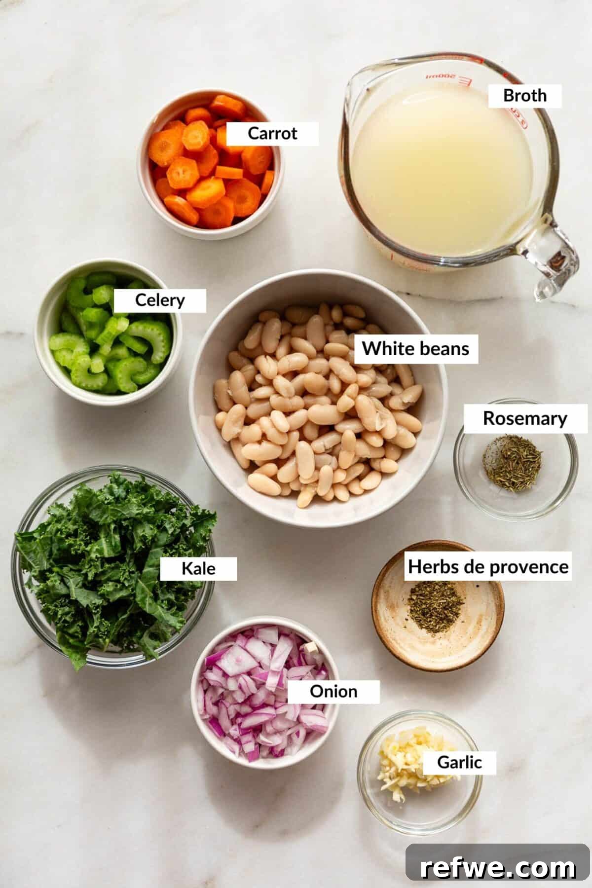 A flat lay of fresh ingredients for Tuscan white bean soup, including cannellini beans, kale, shallots, garlic, carrots, celery, and rosemary.