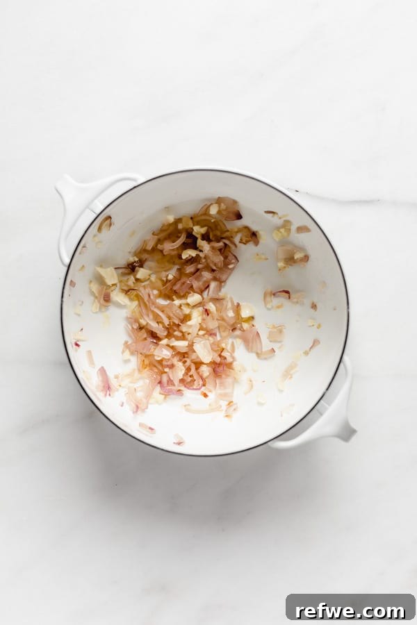 A white pot on a stovetop containing sautéed shallots and garlic, forming the aromatic base of the soup.