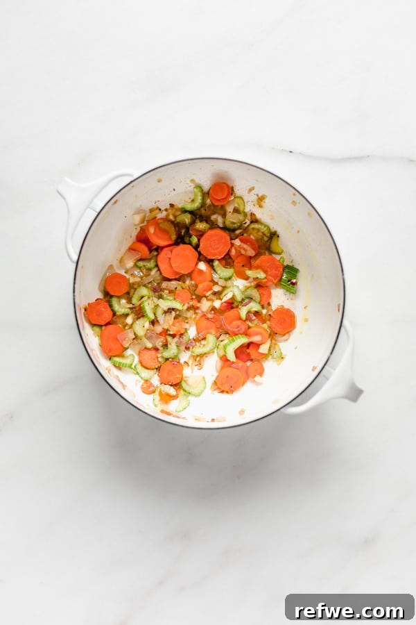 A white pot filled with a colorful mix of chopped carrots, celery, shallots, and garlic, actively sautéing on the stove.
