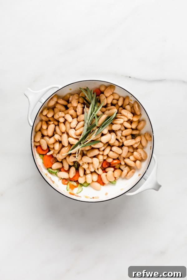 A white pot filled with white beans, herbs, and a sprig of fresh rosemary, awaiting the broth.