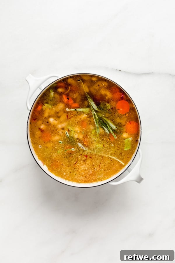 A white pot containing a mix of vegetables, beans, and seasonings, now filled with vegetable broth, simmering on the stove.
