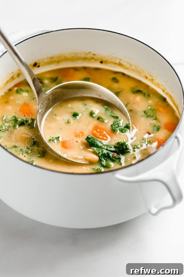 A ladle scooping out a serving of creamy Tuscan white bean and kale soup from a white pot.