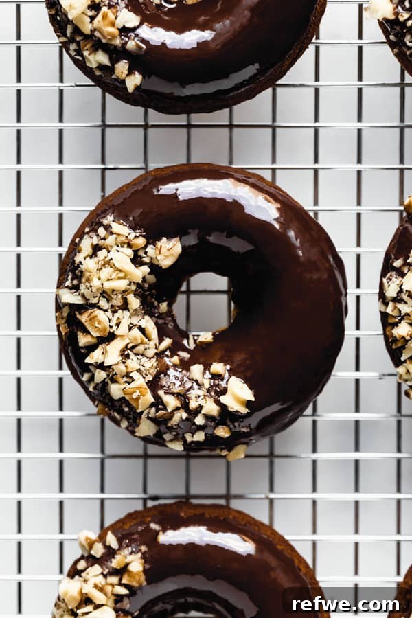 Soft Baked Vegan Banana Bread Donuts 3 A close-up view of a baked vegan banana bread donut, beautifully glazed with chocolate and topped with crunchy crushed walnuts, resting on a silver cooling rack.