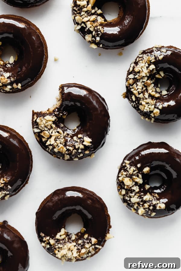 Soft Baked Vegan Banana Bread Donuts 4 A trio of freshly baked vegan banana bread donuts, glistening with chocolate glaze and adorned with crushed walnuts, presented on a clean white background.