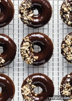 Nine irresistible vegan banana bread donuts, glistening with chocolate ganache glaze and adorned with crushed walnuts, neatly arranged on a silver cooling rack.