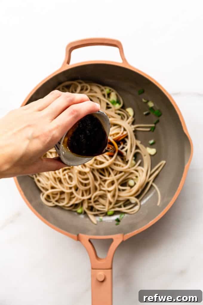 A hand gently pouring the savory stir-fry sauce into a pan of hot noodles.