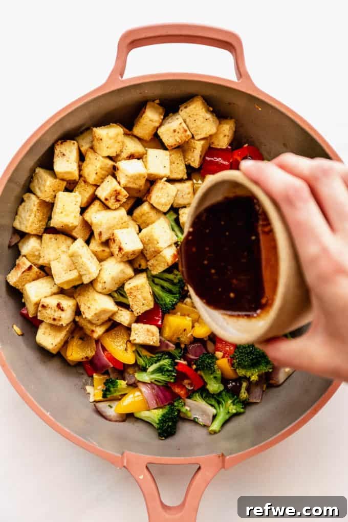 A hand pouring homemade stir fry sauce over a pan filled with crispy tofu and colorful vegetables.