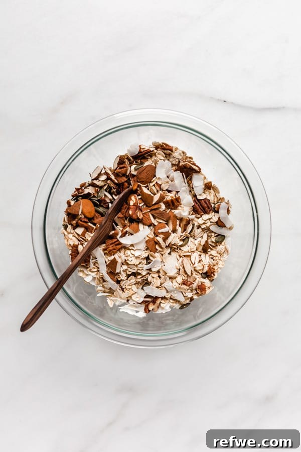 Citrus Cardamom Tahini Granola Crunch 4 A clear mixing bowl filled with large flaked oats, chopped pecans and almonds, pumpkin seeds, coconut flakes, and sesame seeds.