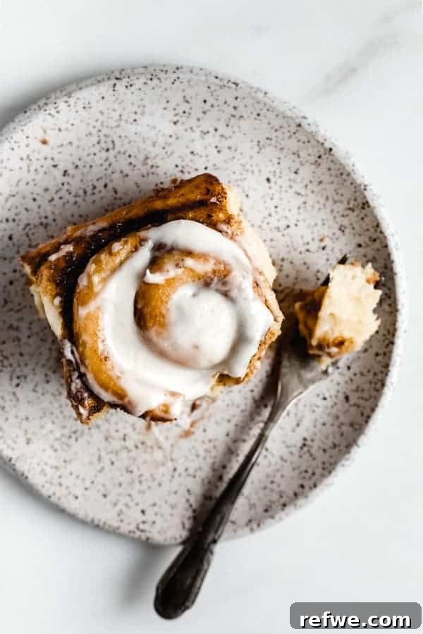 Cozy Vegan Pumpkin Spice Cinnamon Buns 5 A single vegan pumpkin spice cinnamon roll on a white speckled plate, generously topped with smooth cream cheese frosting, ready to be eaten.