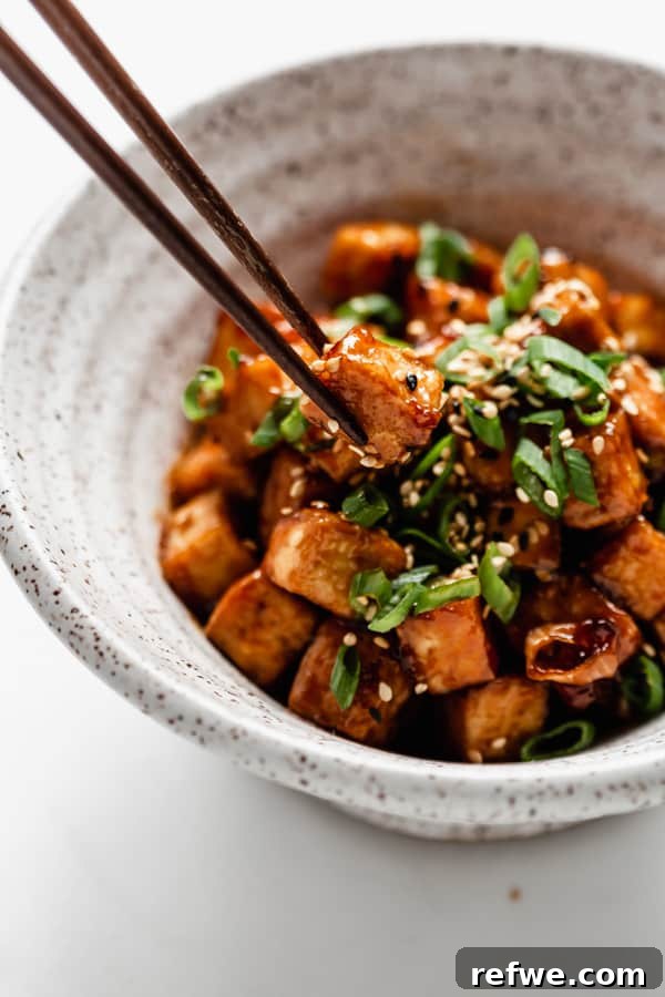 Chopsticks skillfully picking up a perfectly glazed and crispy cube of General Tso Tofu.