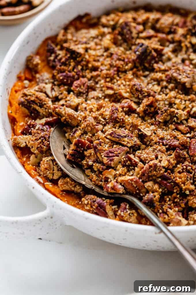 A serving spoon lifting a portion of warm vegan sweet potato casserole with golden pecan crumble from a round dish.