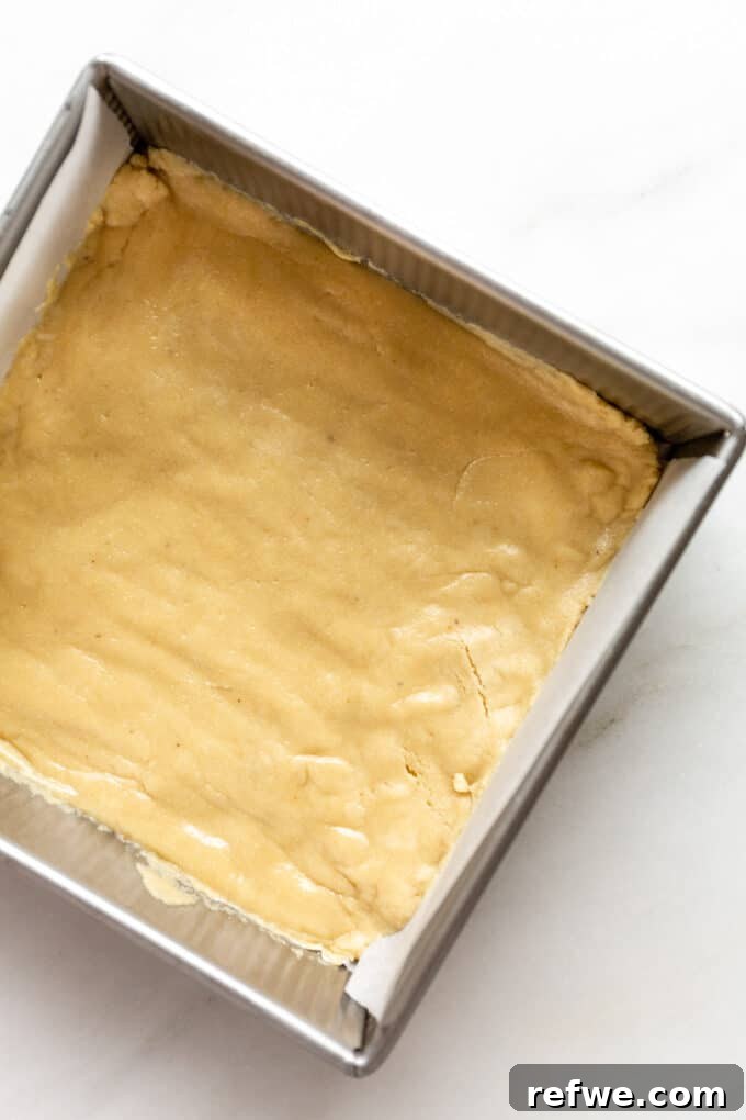 The unbaked almond flour dough is meticulously pressed into an 8x8 inch baking pan, lined with parchment paper, creating a smooth and compact crust base.