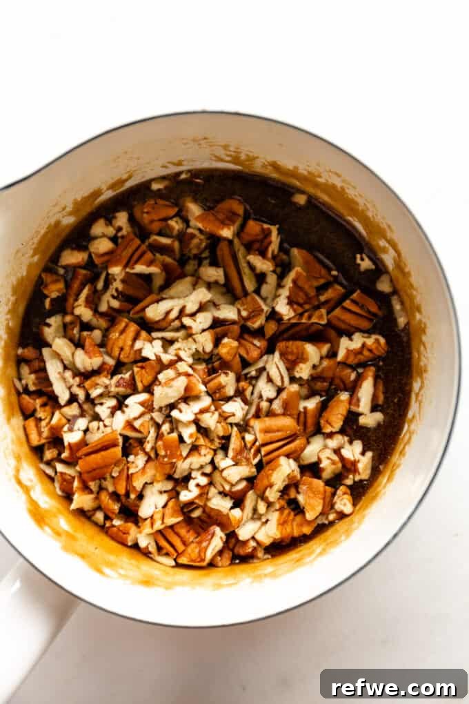 Chopped pecans are gently folded into the warm, gooey caramel mixture in a pot, ensuring each nut is thoroughly coated and glistening before being spread over the crust.