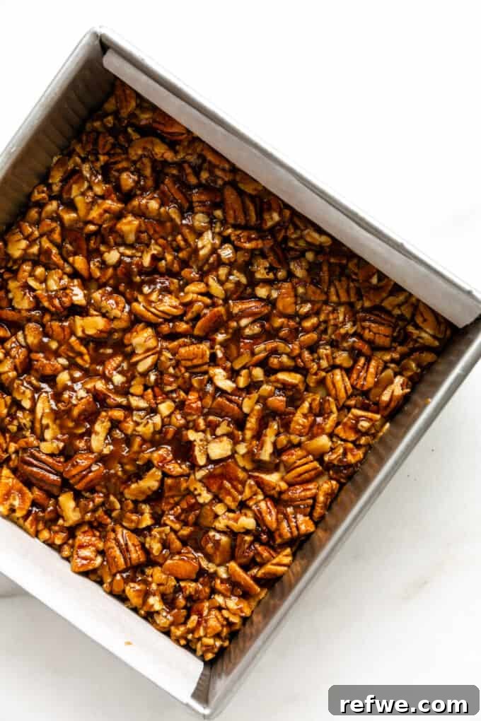 The luscious pecan topping, glistening with caramel, is carefully poured and spread evenly over the golden-brown, pre-baked almond cookie crust in the baking pan.