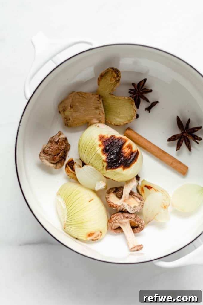 Aromatic Vegan Pho 3 A top-down view of vibrant pho ingredients including white onions, dried shiitake mushrooms, a cinnamon stick, star anise, and fresh ginger, all nestled in a white pot, ready for the first step of broth preparation.