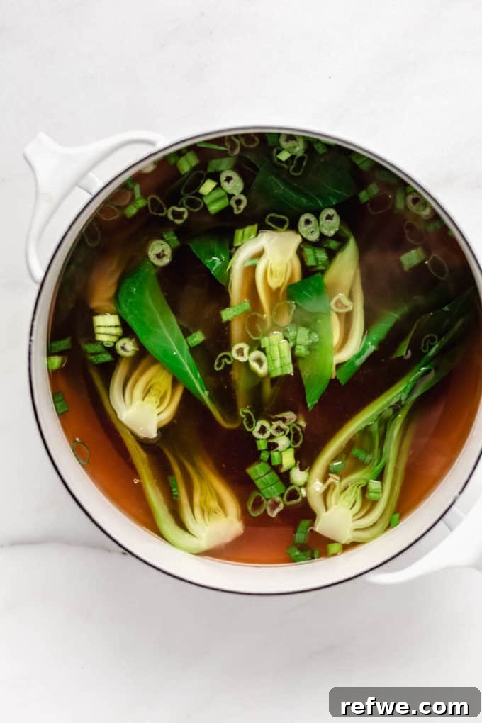 Aromatic Vegan Pho 6 The vibrant green of fresh bok choy and chopped green onions stand out in a white pot filled with vegan pho broth, indicating the final stage of vegetable cooking before serving.