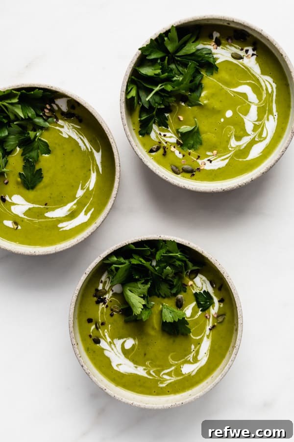 Three beautifully arranged bowls of creamy broccoli soup, each garnished with a swirl of coconut milk and fresh parsley, presented on a wooden surface.