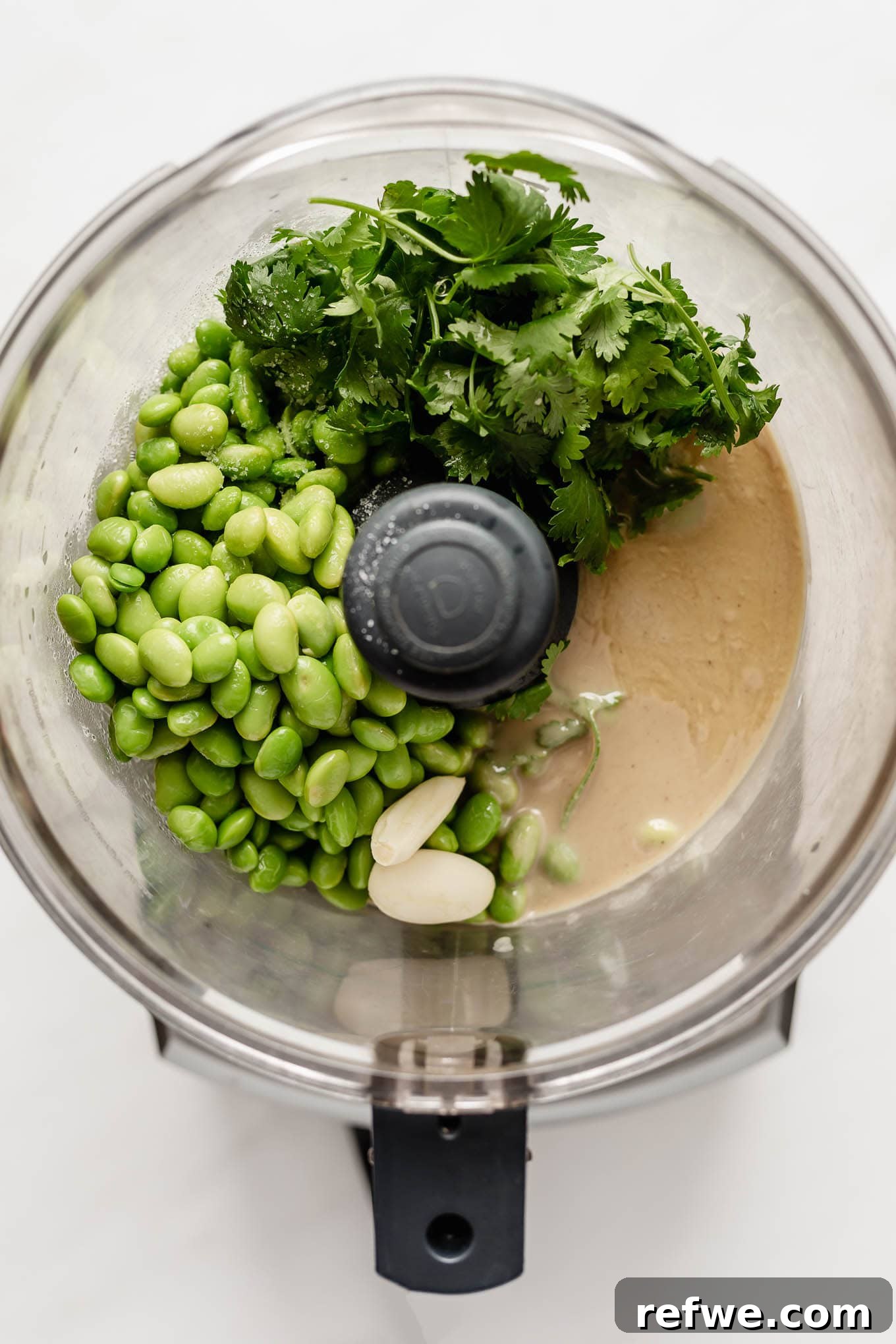 Edamame beans, tahini, fresh herbs, and garlic cloves arranged in a food processor bowl before blending.
