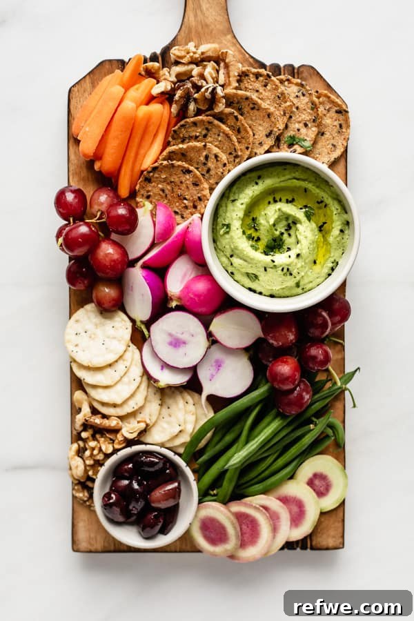 A beautifully arranged wooden platter featuring a bowl of edamame hummus surrounded by fresh veggies, fruits, and crackers.