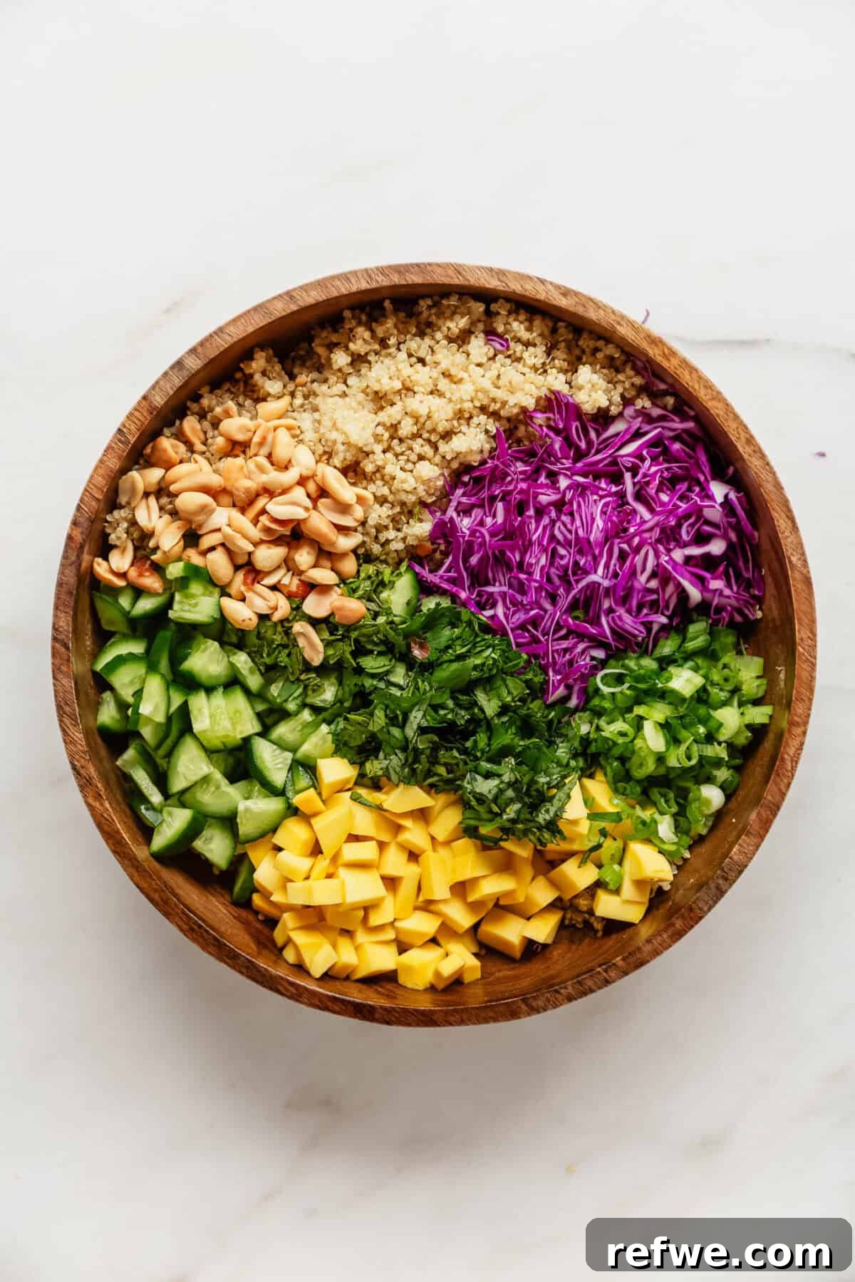 Thai Peanut Quinoa Bowl 6 All the crisp and colorful ingredients for the Thai quinoa salad, including cooked quinoa, shredded red cabbage, diced cucumber, fresh mango cubes, and chopped herbs, are neatly arranged in a spacious wood salad bowl, ready for the dressing.