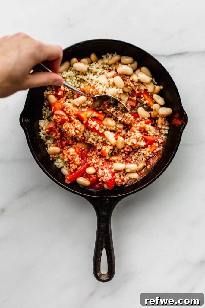 Hearty Vegan Shakshuka 5 A hand gently mixing wholesome quinoa and creamy white beans into the rich tomato and pepper sauce within a cast iron skillet, ensuring even distribution of flavors.