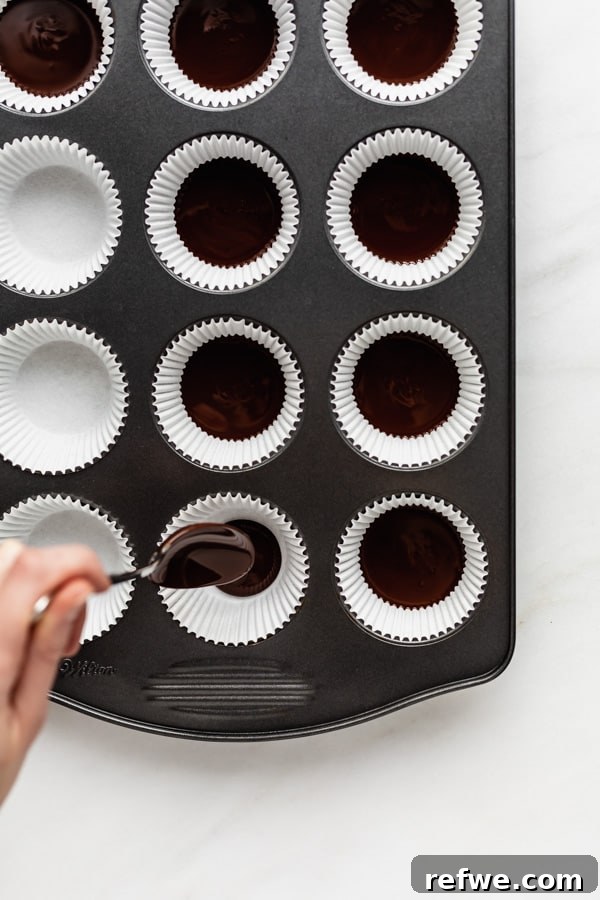 Decadent Dark Chocolate Almond Butter Delights 4 A hand spooning the initial layer of melted chocolate into muffin liners in a baking pan.