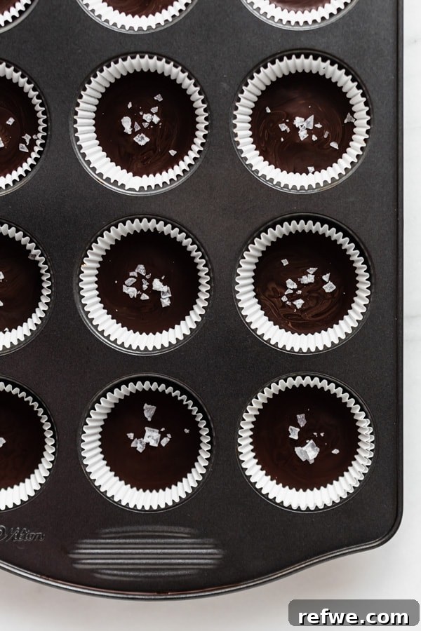 Decadent Dark Chocolate Almond Butter Delights 6 Dark chocolate almond butter cups in a muffin pan, freshly topped with the final chocolate layer and a sprinkle of flaky sea salt.