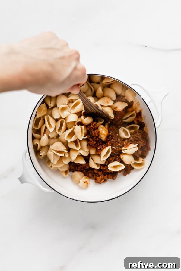 Zesty Plant-Based Taco Pasta 4 A hand expertly mixing the rich vegan taco sauce with shell noodles in a pristine white pot, creating a harmonious blend of flavors.