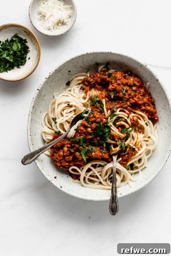 Effortless Elegance: 20 Stunning Date Night Dinners 12 Two silver spoons in a bowl of spaghetti pasta generously topped with rich lentil bolognese and fresh herbs, alongside pinch bowls of extra herbs and cheese on a white table.
