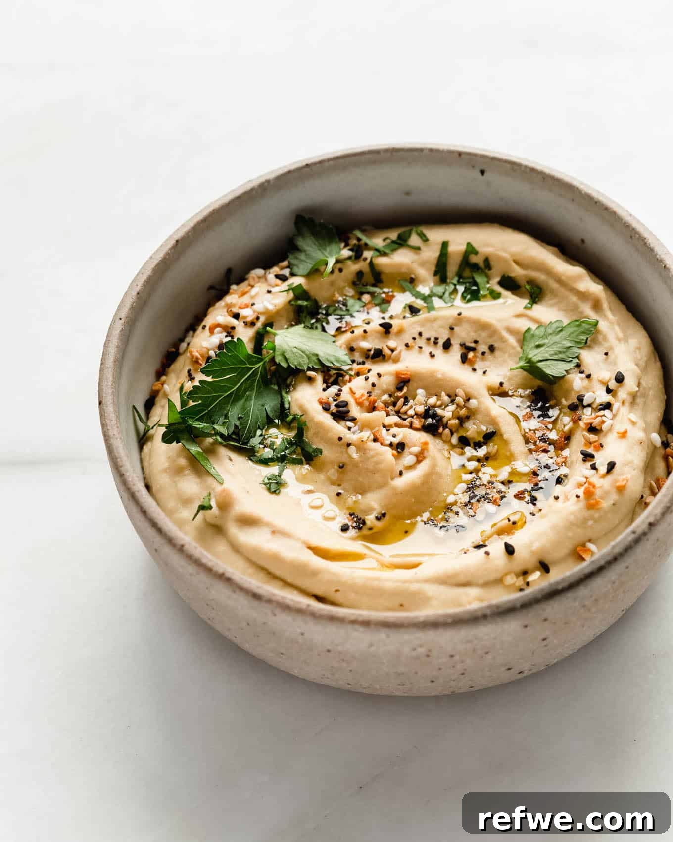 Everything Bagel Seasoned Hummus 6 everything bagel hummus in a bowl topped with herbs and olive oil.