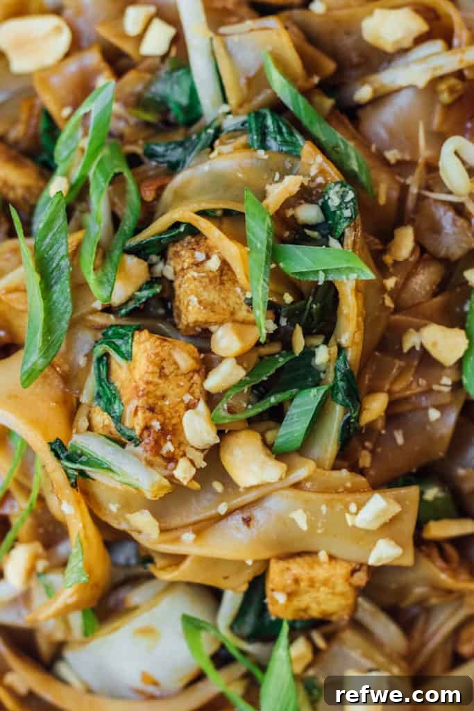 Hearty Vegan Pad See Ew 3 A close-up view of glistening Pad See Ew noodles, perfectly coated in a dark, savory sauce, interspersed with tender Chinese broccoli and crispy tofu pieces.