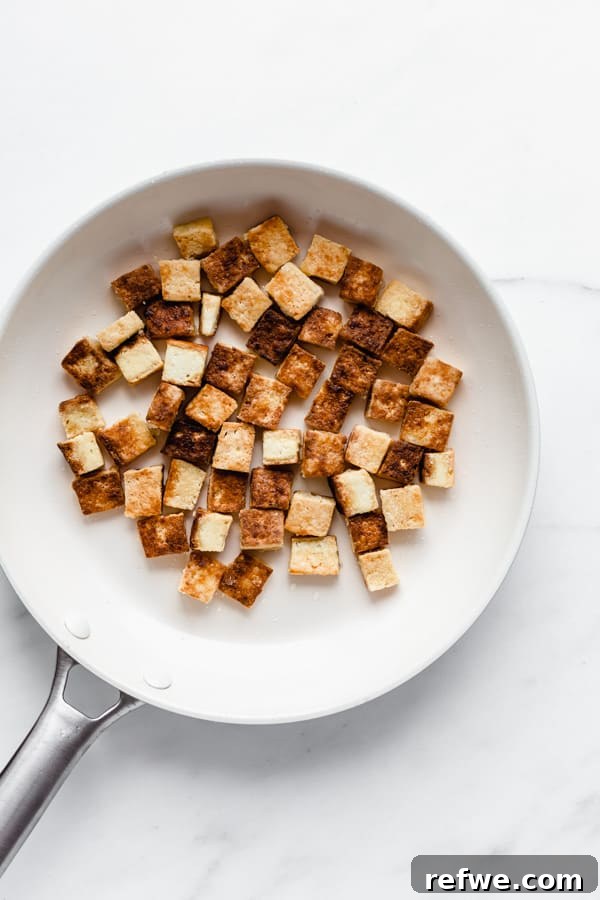 Crispy golden brown tofu cubes cooking in a white non-stick pan.